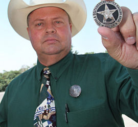 A man in a dress shirt with a light colored cowboy hat one holding up a silver and black circle star badge