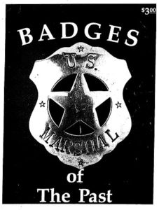 A silver shield shaped badge with the word "U.S" across the top and "Marshal" across the bottom. There are small stars at the top, bottom, and left and right sides of the badge. On the black background of the photograph are the words "Badges of The Past".