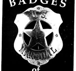 A silver shield shaped badge with the word "U.S" across the top and "Marshal" across the bottom. There are small stars at the top, bottom, and left and right sides of the badge. On the black background of the photograph are the words "Badges of The Past".