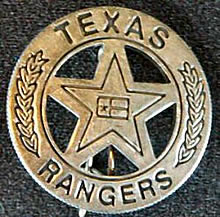 A silver circle star badge with the Texas Flag in the middle of the star, branches on the sides of the circle, and the words "Texas Ranger" on the top and bottom.