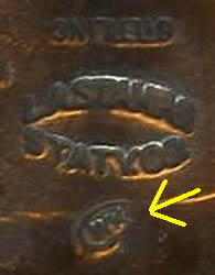 A blurry close up of the back of a bronze badge with unclear writing and a digitally drawn on yellow arrow pointing to a symbol underneath the words.