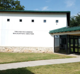A white building with a green roof and a tan stone rim around the bottom. Written on the building are the words "Texas Rangers Education Center".