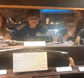 Three children photographed behind an exhibit about decorative firearms.