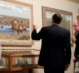 Group of men in dark suits looking at paintings hung on a wall.