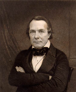 A black and white waist up photograph of a clean shaven man in a dark suit with his arms crossed.