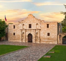 A stone building in the Spanish mission style with a large stone and grass courtyard flying a Texas flag against the setting or rising sun.
