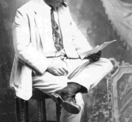 A man in a white cowboy hat and a light colored suit sitting on a stool, reading a paper, and smoking a cigar.