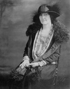 A woman in a formal 1910s gown and fancy hat posing for a black and white photograph.