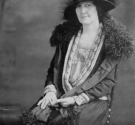 A woman in a formal 1910s gown and fancy hat posing for a black and white photograph.