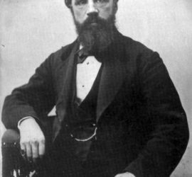 A waist up picture of a man in a dark suit with a beard and a pinkie ring.