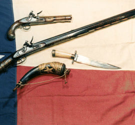 A pistol, an inlaid rifle, bowie knife, decorated powder horn arranged across a Texas flag.