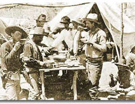 Nine men in 1870s Ranger gear at a campsite eating a meal. Some are standing and some are seated on the ground.