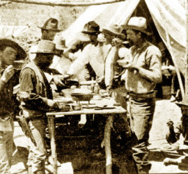 Nine men in 1870s Ranger gear at a campsite eating a meal. Some are standing and some are seated on the ground.