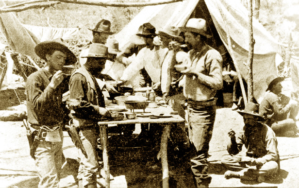 Nine men in 1870s Ranger gear at a campsite eating a meal. Some are standing and some are seated on the ground.