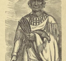 A Tonkawa man in fringed regalia holding a pipe, a dead bird, and a handful of feathers.