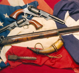 Two pistols, a rifle, a powder horn, and another tool arranged across an American and Texas flag.