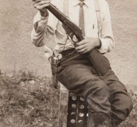A 1900s Ranger in a collared shirt, tie, cowboy boots, and a cowboy hat poses for a picture with his rifle.