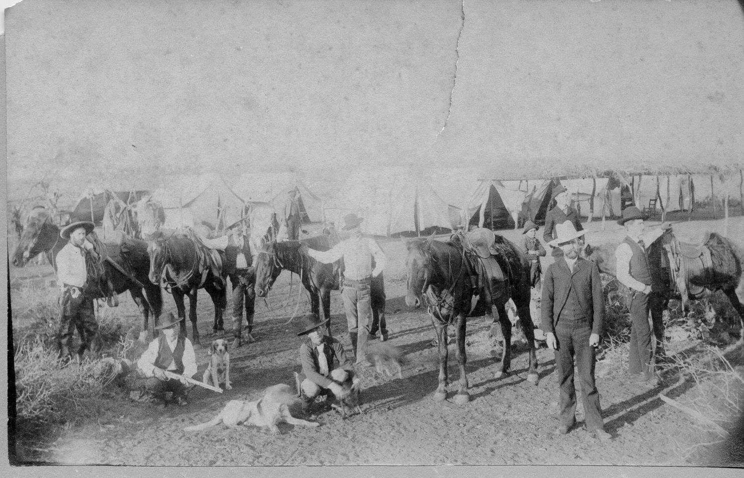 Multiple men, horses, and dogs in front of the many tents that made up a Ranger camp in the 18070s.