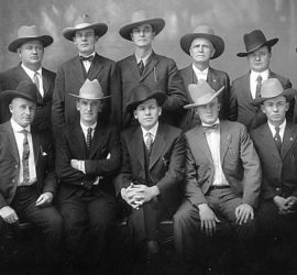 Twelve men posing for a picture in 1920s dark suits and cowboy hats.