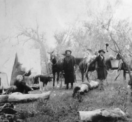 An 1890s Ranger campsite with many men and horses and one dog spread out. One man is playing the banjo and all of the men are wearing hats.