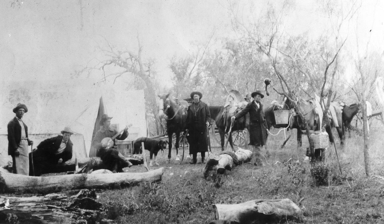 An 1890s Ranger campsite with many men and horses and one dog spread out. One man is playing the banjo and all of the men are wearing hats.