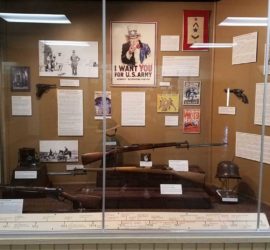An exhibit with multiple firearms, a US Army poster, helmets, and photographs of World War One.