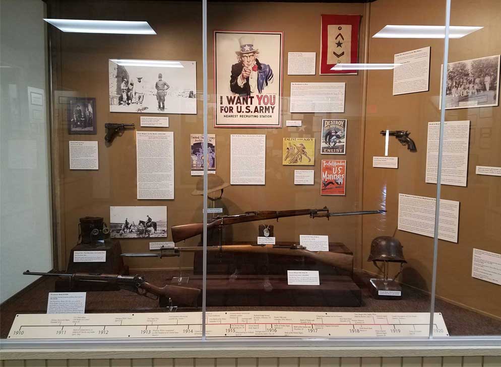 An exhibit with multiple firearms, a US Army poster, helmets, and photographs of World War One.