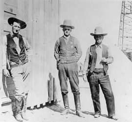 Three early 1900s Rangers in vests and cowboy hats standing in front of a building with an oil rig in the background.