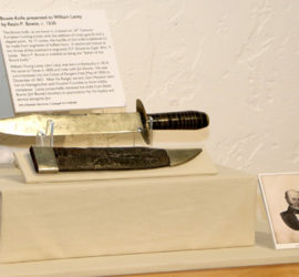 A bowie knife and a leather a silver metal scabbard on exhibit with a label and an old picture of a man in a suit.