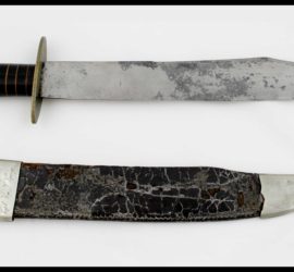 A silver metal bowie knife with a black and bronze colored handle above a silver metal and black leather scabbard with engraving on the top.
