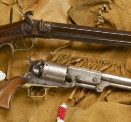 A rifle with an intricate brass trigger guard and pistol with wooden grips arranged across a fringed and beaded suede jacket.