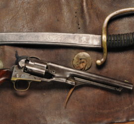 A Colt pistol with wooden grips below a cavalry saber with a brass handguard. Both are sitting on a pair of dark brown leather chaps with silver circular decorations.