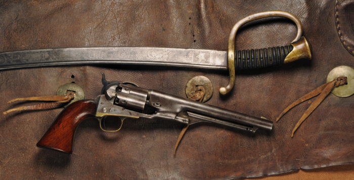 A Colt pistol with wooden grips below a cavalry saber with a brass handguard. Both are sitting on a pair of dark brown leather chaps with silver circular decorations.