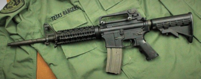 A dark grey assault rifle against a green uniform shirt with "Texas Rangers" embroidered above the right pocket.