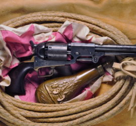 A dragoon revolver pistol next to a bronze black powder container with an engraving of a cannon, a flag, and various spears. Both are nestled in a pink and white cloth in a coiled braided whip or rope.