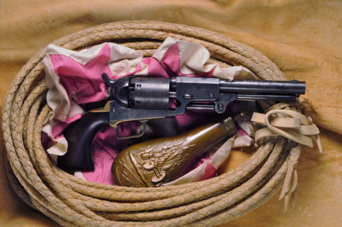 A dragoon revolver pistol next to a bronze black powder container with an engraving of a cannon, a flag, and various spears. Both are nestled in a pink and white cloth in a coiled braided whip or rope.