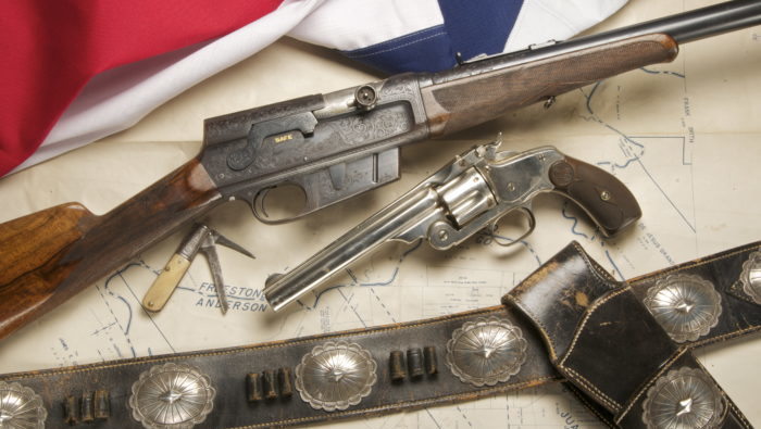 One rifle, one pistol, a pocket knife, and a black gunbelt with silver accents arrange across an older map. The edge of the Texas flag is visible in the top left corner.