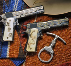 Two highly decorated automatic pistols next to a single handcuff with the brim of a tan hat visible in the edge. Both firearms are a dark silver metal with gold decorations and ivory grips. One grip has the golden initials "MTG" and the other has an engraved long horn with golden horns. Everything is arranged on a multicolored saddle blanket.