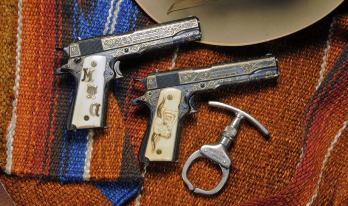 Two highly decorated automatic pistols next to a single handcuff with the brim of a tan hat visible in the edge. Both firearms are a dark silver metal with gold decorations and ivory grips. One grip has the golden initials "MTG" and the other has an engraved long horn with golden horns. Everything is arranged on a multicolored saddle blanket.