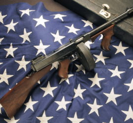 Thompson Submachine gun with dark grey metal and darker wooden areas arranged on the star section of an American flag.