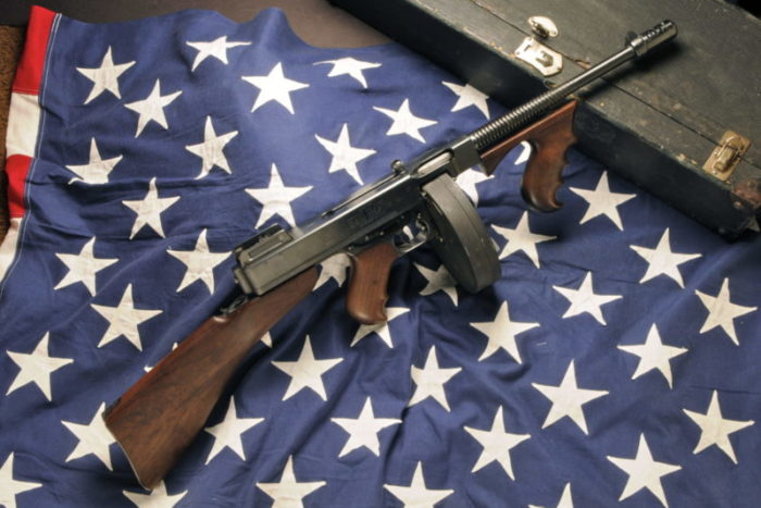 Thompson Submachine gun with dark grey metal and darker wooden areas arranged on the star section of an American flag.