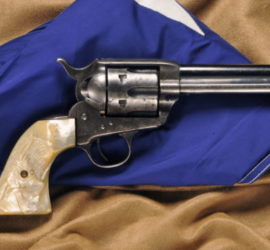 A silver metal pistol with mother of pearl grips engraved with a long horn. The firearm is resting against a folded up Texas flag.