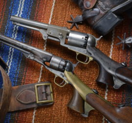 An older leather belt, two Colt pistols with attached shoulder stocks, and the heels with spurs of a pair of boots all positioned on a multicolored saddle blanket.