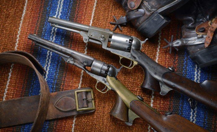 An older leather belt, two Colt pistols with attached shoulder stocks, and the heels with spurs of a pair of boots all positioned on a multicolored saddle blanket.