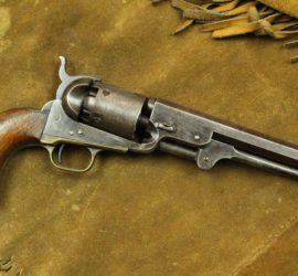 A silver metal pistol with a wooden butt on top of a fringed suede background.