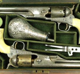 A box lined in green velvet with two silver pistols, a silver black powder container, two silver accessories, and two jars holding caps.