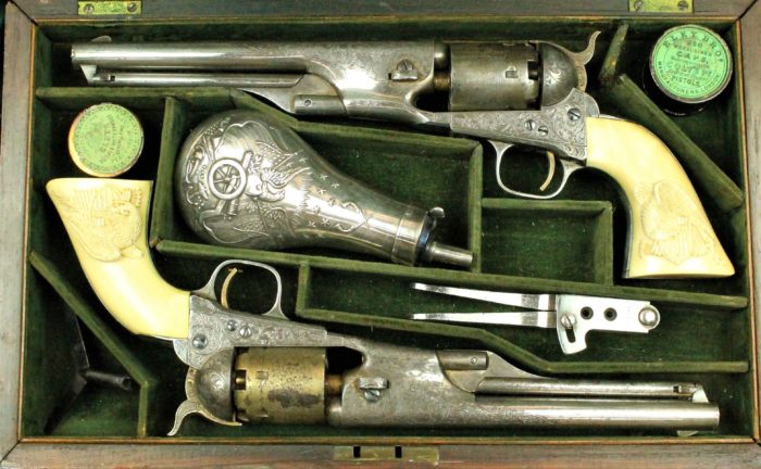 A box lined in green velvet with two silver pistols, a silver black powder container, two silver accessories, and two jars holding caps.