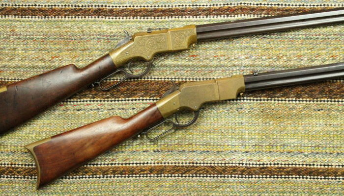 Two repeating rifles with wooden butts and brass receivers. One gun's receiver has engraving on it. Both guns rest on a multicolored saddle blanket.