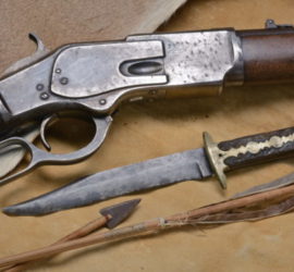 The middle part of a rifle, a bowie knife with an inlaid handle, and two arrows arrange on the tanned side of a fur.