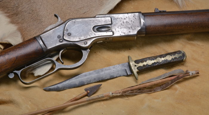 The middle part of a rifle, a bowie knife with an inlaid handle, and two arrows arrange on the tanned side of a fur.